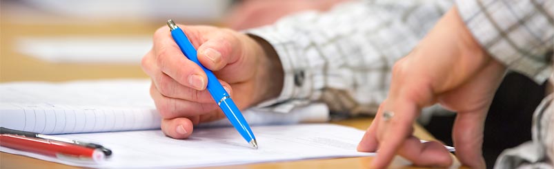 En hand med en bläckpenna och linjerat papper på ett bord.