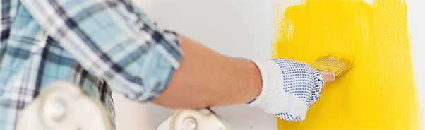 Foto på en målare som målar en vägg inomhus med pensel och gul målarfärg.